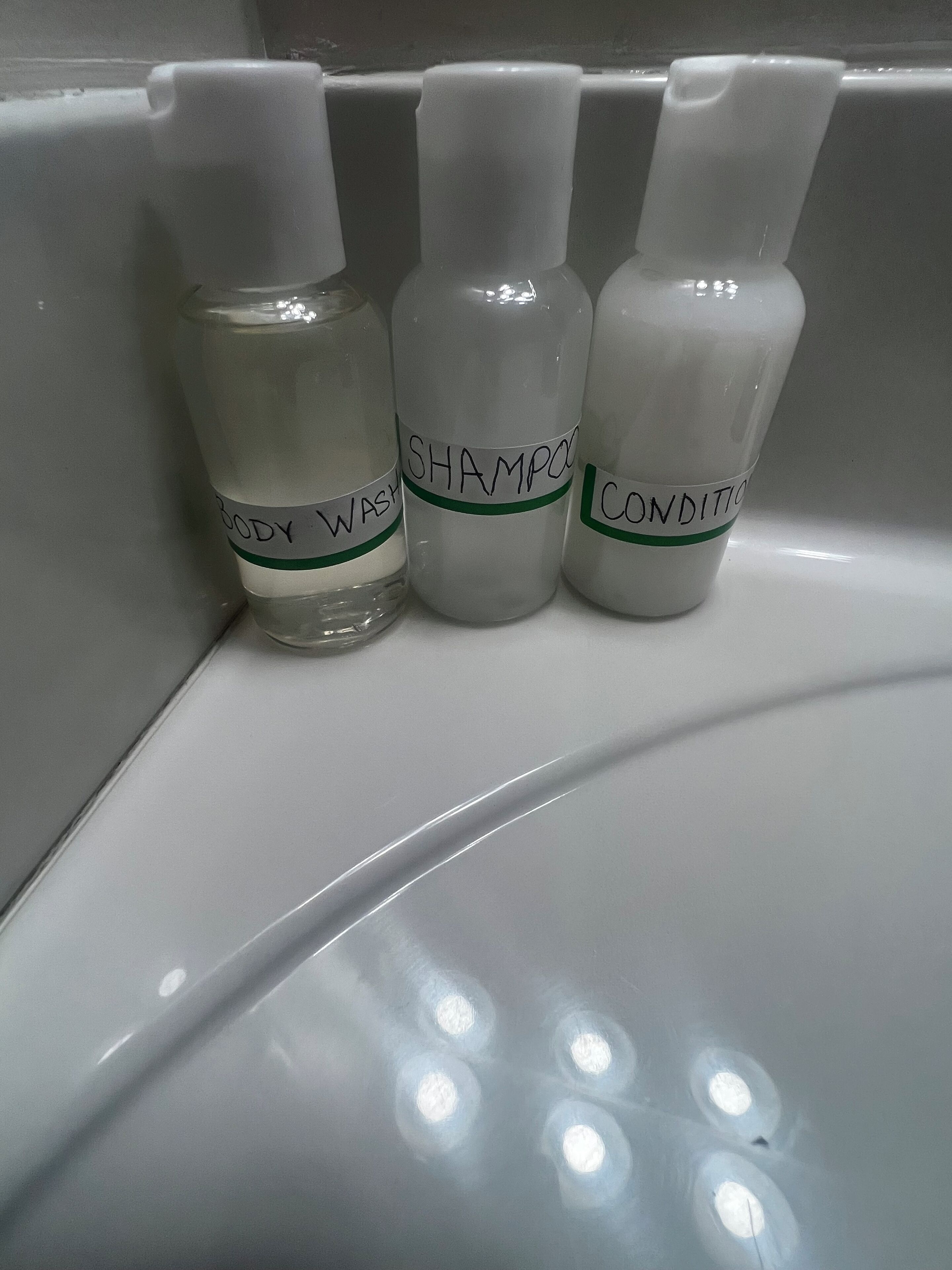Three bottles labeled body wash, shampoo, and conditioner on a white shelf.