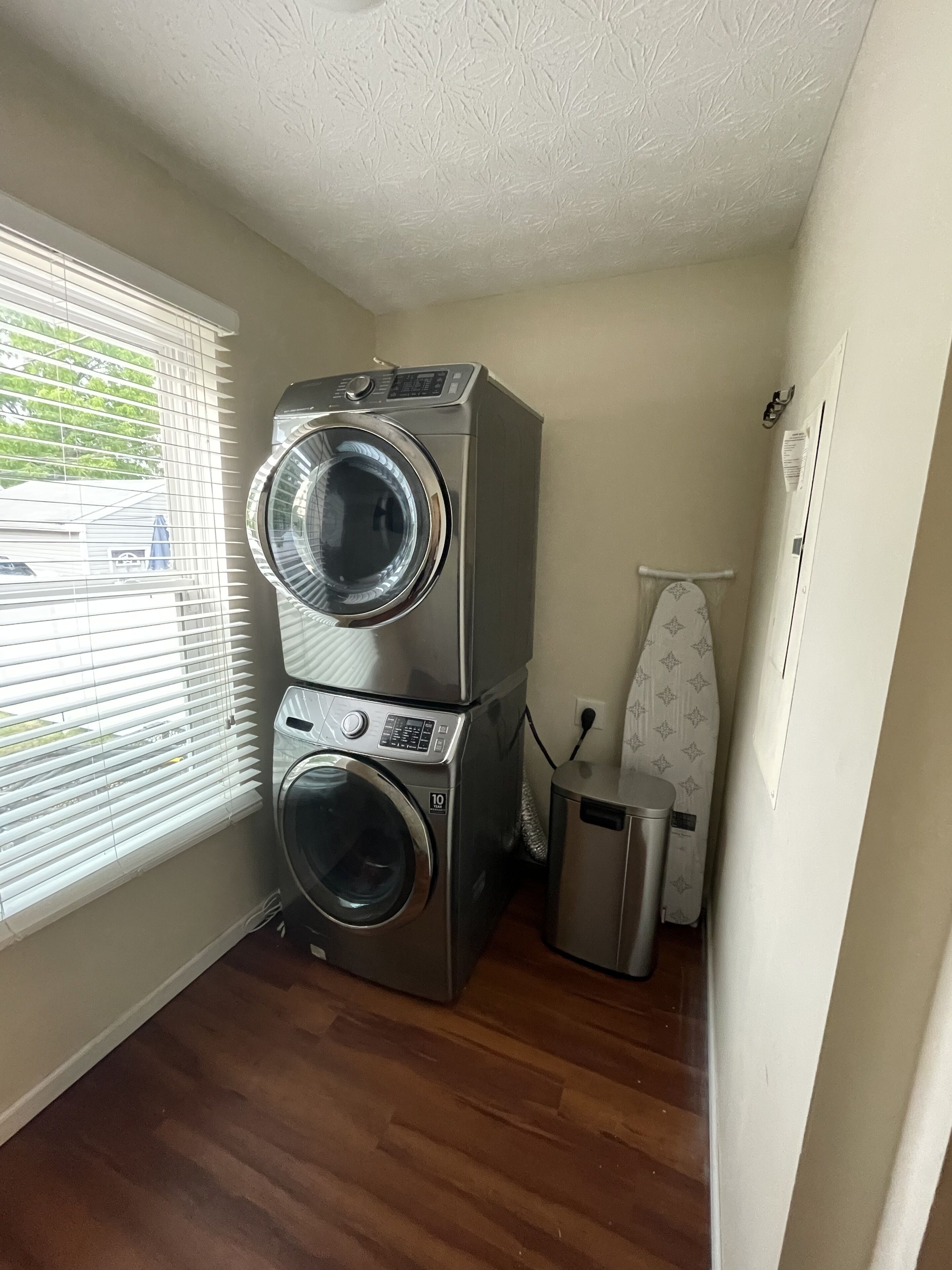 A laundry room with two stacked washing machines, a small trash bin, and an ironing board.