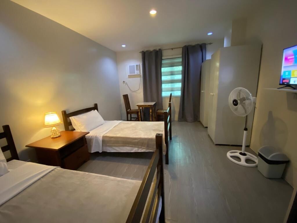 Habitación de hotel con dos camas, un escritorio, una silla, un televisor, un ventilador y una ventana con cortinas.