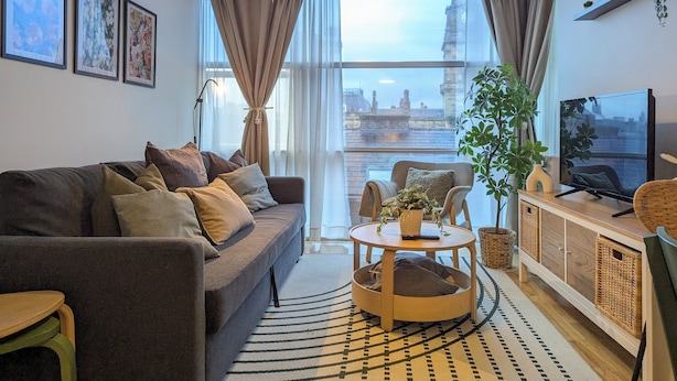 Ein modernes Wohnzimmer mit einer Couch, einem runden Couchtisch, einem Fernseher und Blick auf eine Stadtlandschaft durch große Fenster.