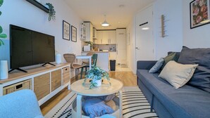 Ein modernes Wohnzimmer mit grauem Sofa, einem Fernseher auf einem Holzbodenständer, einem kleinen runden Tisch mit einer Pflanze und einer Küche im Hintergrund.