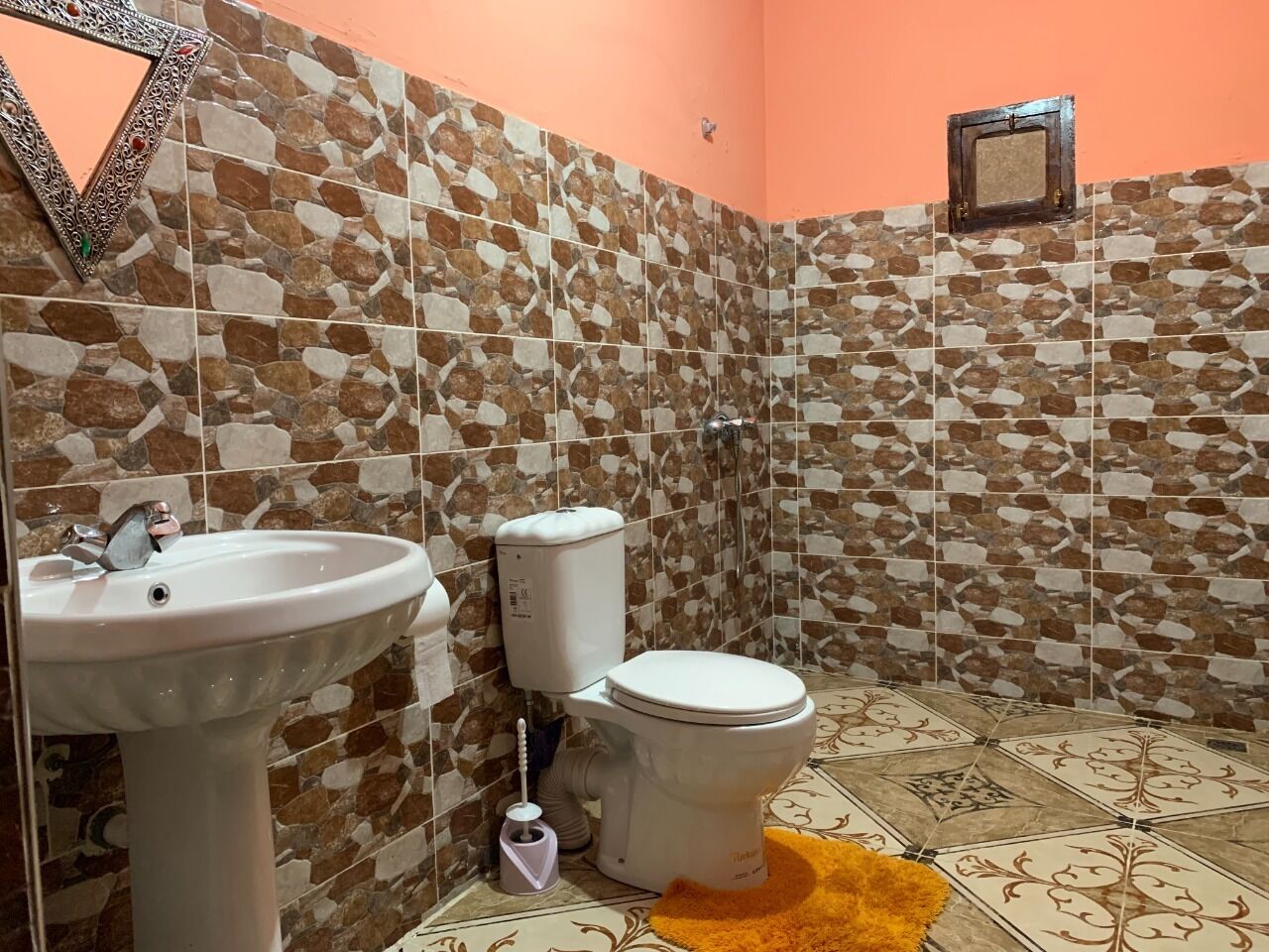 Ein Badezimmer mit einem weißen Waschbecken, einer Toilette und einer Dusche. Die Wände sind mit einem braun-beigen Muster gefliest.