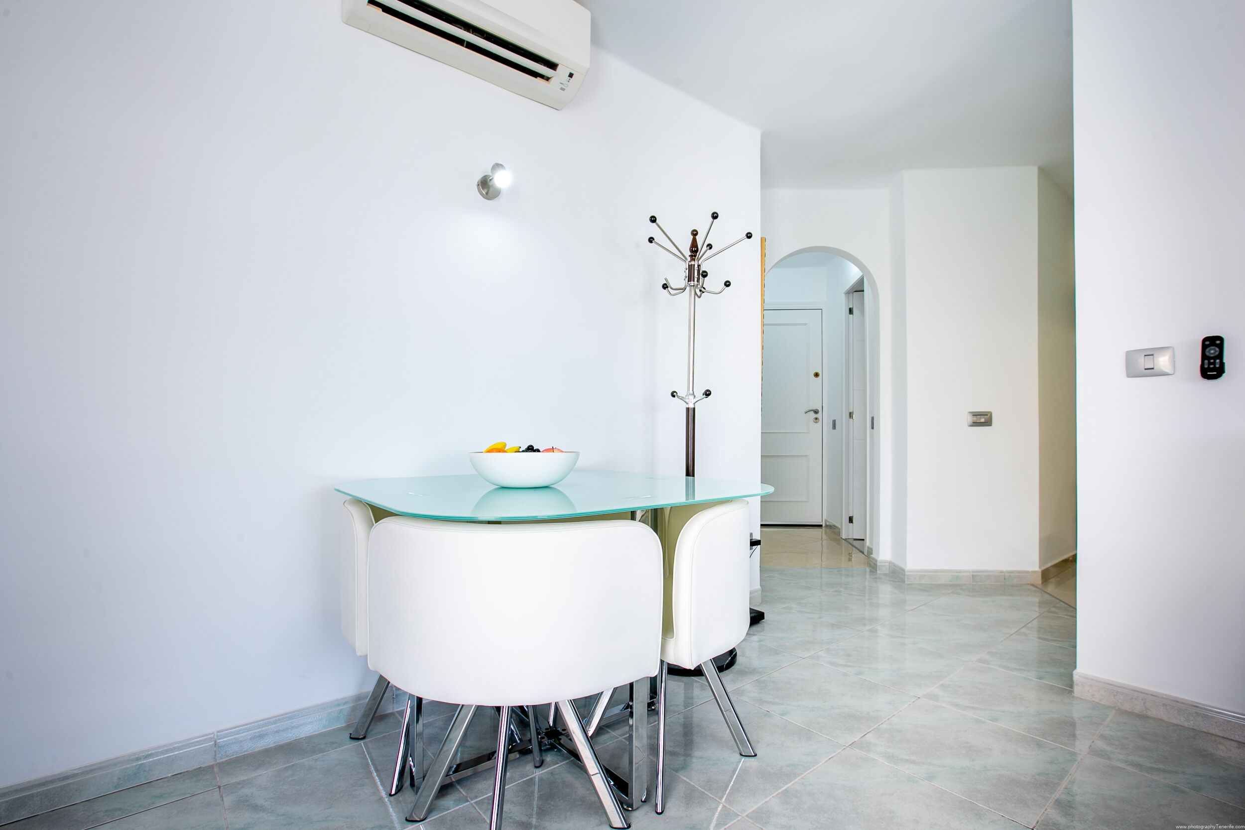 A modern dining area with a glass table, white chairs, and a bowl of fruit.