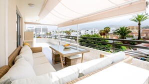 A balcony with wicker furniture, a glass table, and a view of buildings and greenery.