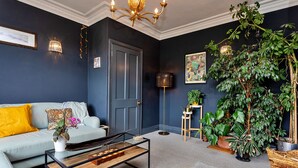 A modern living room with a dark blue wall, a light grey sofa, a glass coffee table, and a variety of plants.