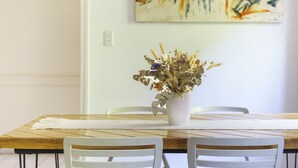 Una mesa de comedor con un jarrón blanco de flores secas, sillas blancas y un cuadro en la pared.