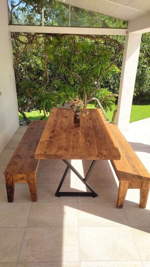 Mesa de madeira com bancos sob um espaço coberto, com vista para o verde.