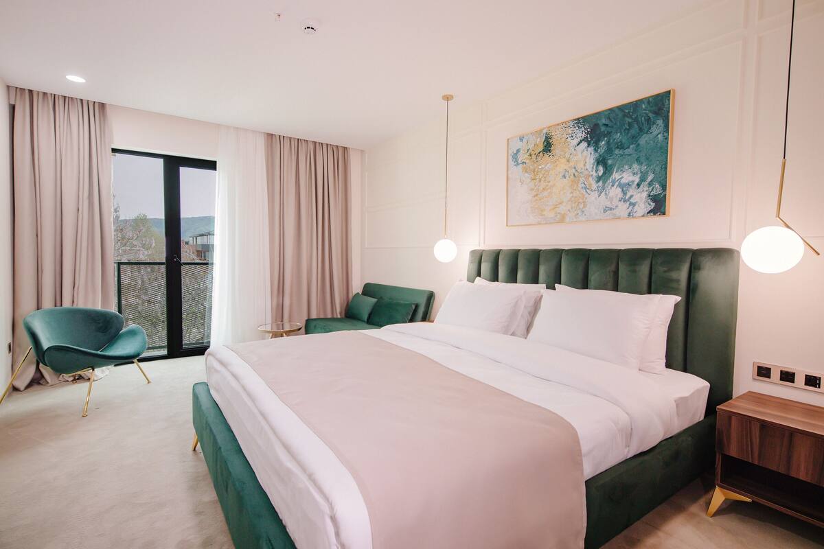 A modern hotel room with a large bed, a green armchair, a small table, and a view of the outdoors through a sliding door.