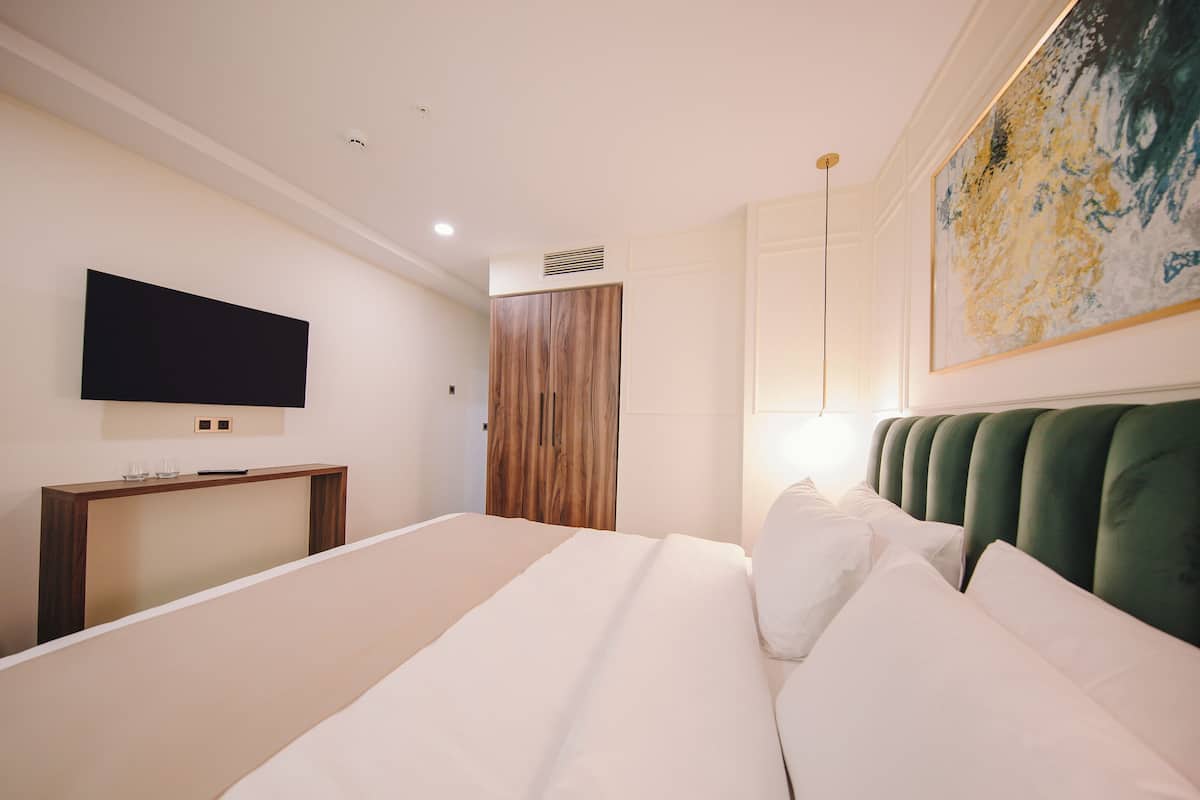 A modern hotel room with a large bed, a flat-screen TV mounted on the wall, a wooden wardrobe, and a green upholstered headboard.