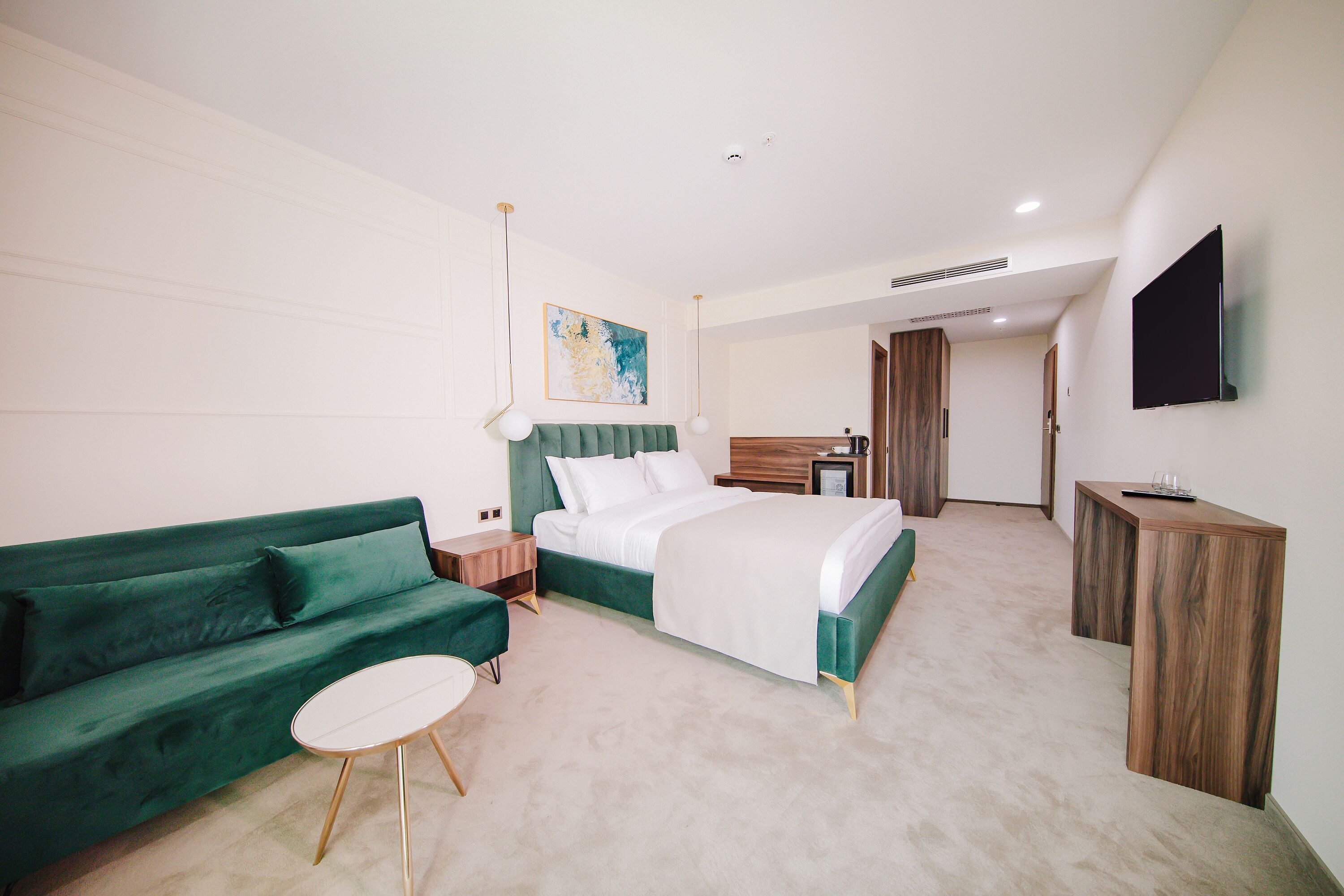 A modern hotel room with a green sofa, a single bed, a small round table, a wooden desk, and a flat-screen TV mounted on the wall.