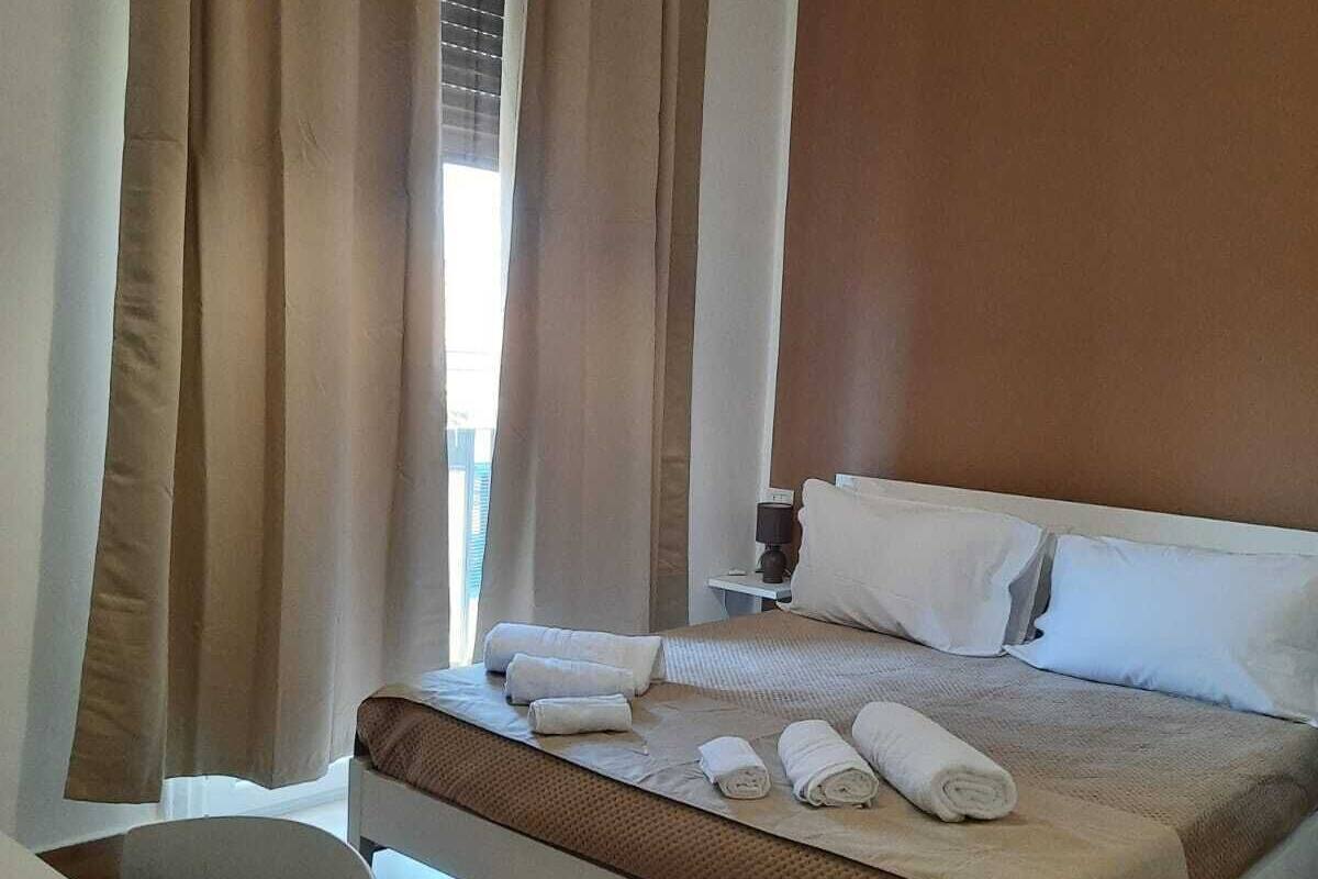 Quarto de hotel com cama, mesa de cabeceira, luminária, ar condicionado e vista para o exterior pela janela.