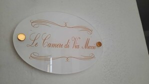 A white, oval-shaped sign with gold lettering and decorative swirls, featuring the text "Le Cameri di Via Meccio." - Via Salvatore Meccio (Palermo)