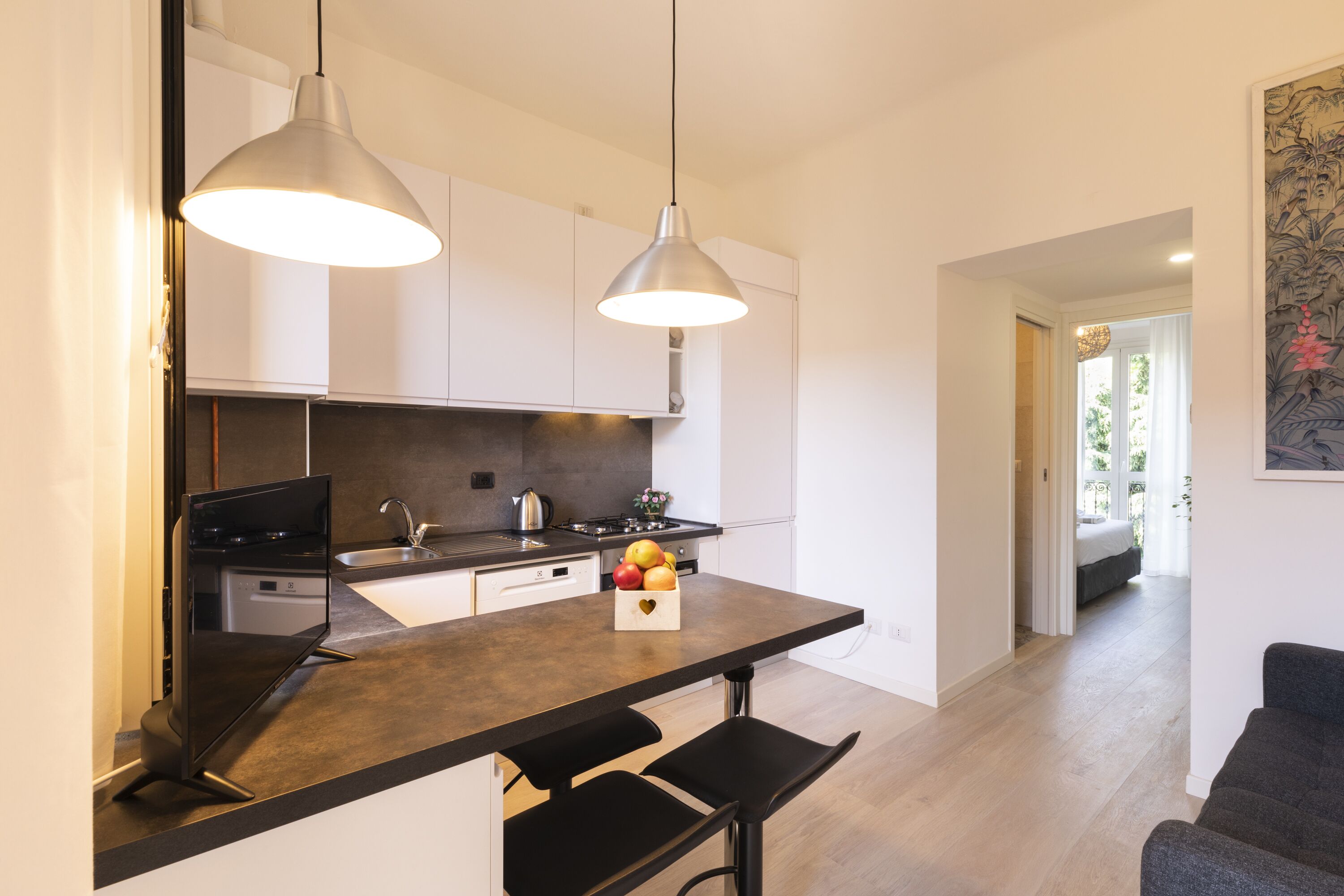 A modern kitchen with a dark countertop, two pendant lights, a fruit basket, and a view into the living area.