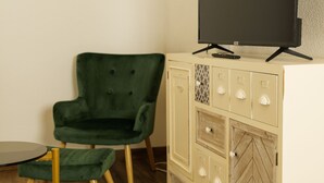 Een kamer met een groene fauteuil, een klein rond tafeltje, een wit bergkastje met lades en een flatscreen televisie aan de muur.