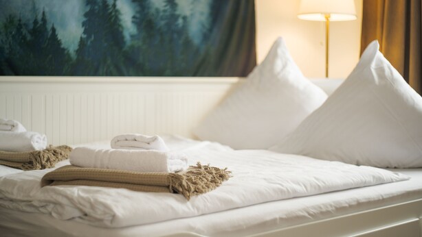 Een net opgemaakt bed met witte lakens, een beige plaid en twee witte kussens. Op de muur achter het bed is een schilderij van een bos te zien.