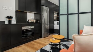 A modern kitchen with black cabinets, a stainless steel refrigerator, and a built-in oven. There is a small wooden table and a chair with orange cushions. - Vibrant Central Studio at The Carrington (Cape Town)