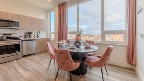 A modern kitchen with stainless steel appliances, a dining area with pink upholstered chairs, and large windows offering a city view.