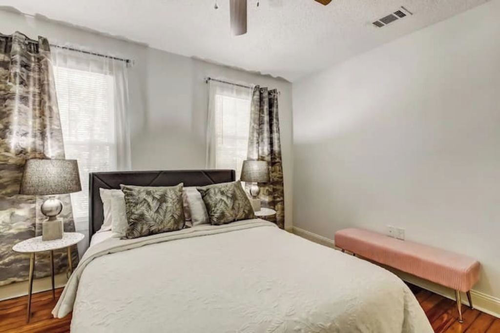 Une chambre à coucher avec un lit, deux tables de chevet avec des lampes, un banc et deux fenêtres avec des rideaux.