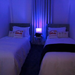 Two single beds with white bedding and pillows, a table lamp, and a potted plant.