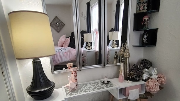 A bedroom with a vanity, a large mirror, a bedside table, a lamp, a small trash bin, and a white ottoman.