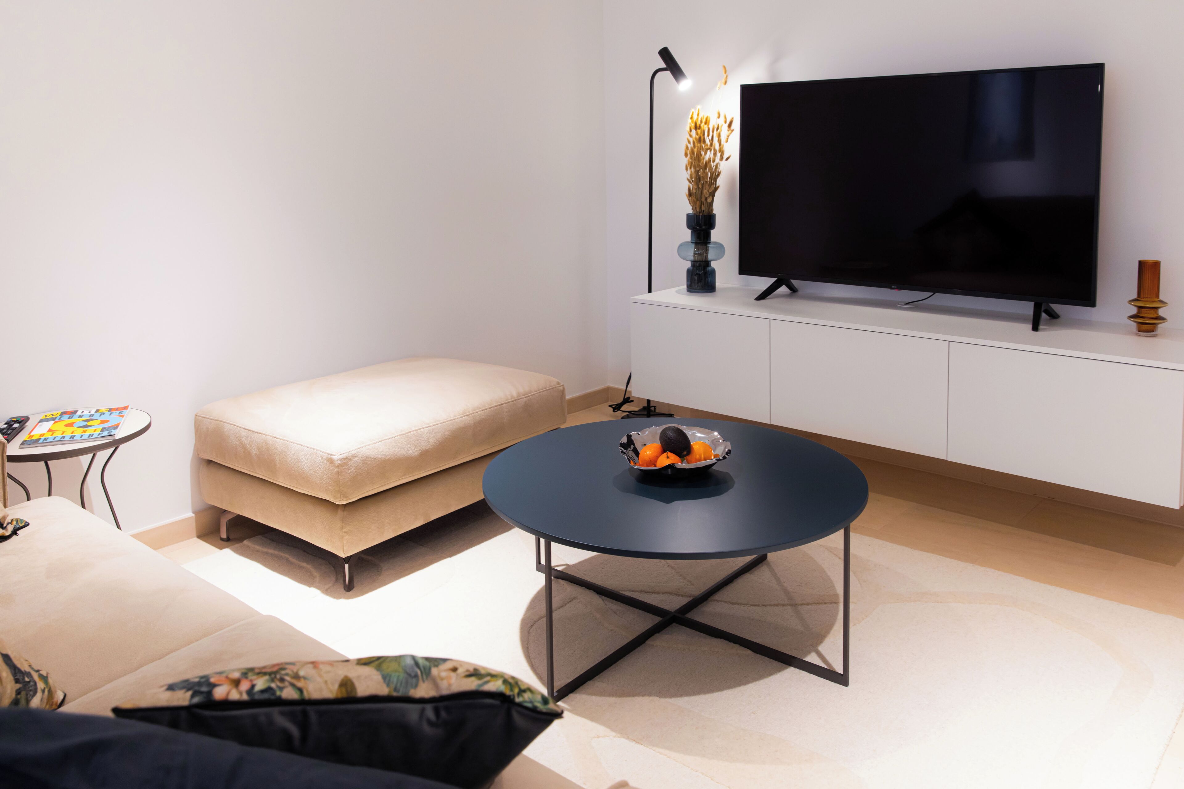Un salón moderno con un televisor de pantalla plana, una mesa de centro redonda con naranjas, un sofá color beige y una mesita auxiliar con una revista.