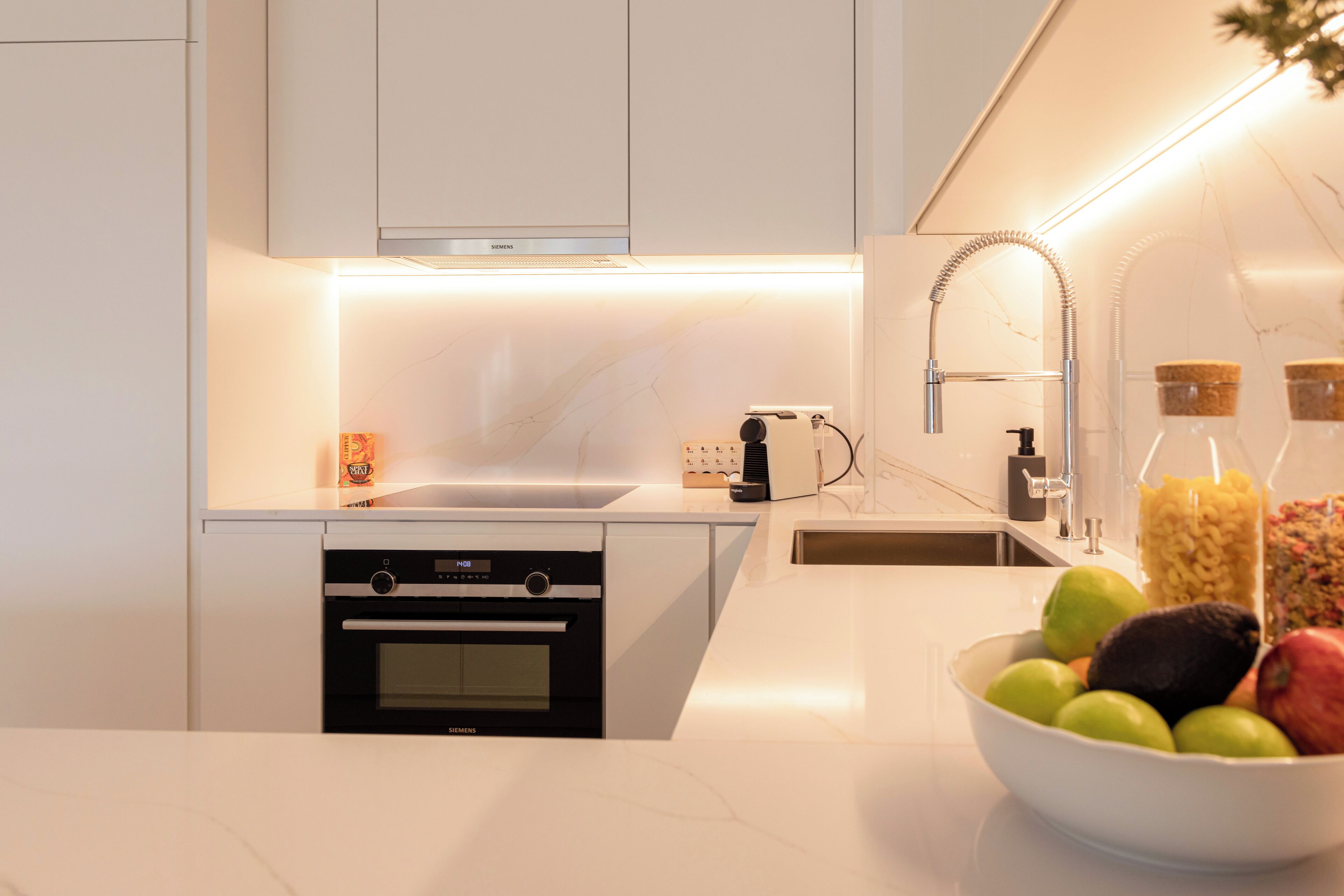 Una cocina moderna con armarios blancos, horno empotrado, lavamanos y un cuenco de fruta sobre la encimera.
