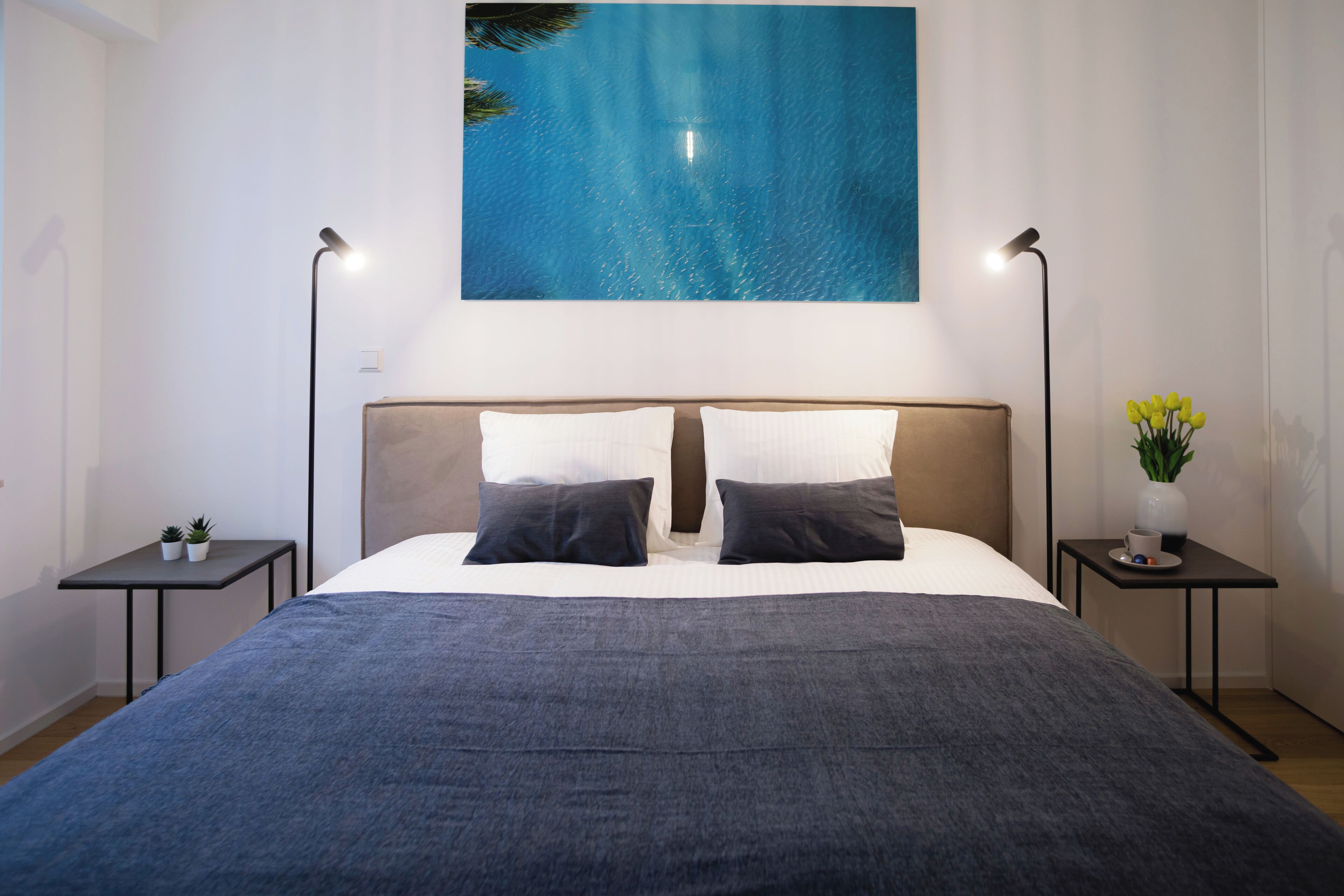 Un dormitorio ordenado con una cama grande, dos mesitas de noche, una mesita auxiliar, un cuadro colgado en la pared y una pequeña planta en maceta.