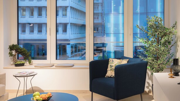 Un salon moderne avec un fauteuil bleu foncé, une table basse ronde surmontée d’un panier de fruits, une petite table d’appoint avec un magazine, et de grandes fenêtres donnant sur un immeuble doté de balcons.