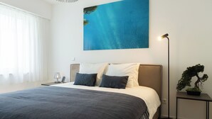 Une chambre moderne avec un grand lit, une table de chevet avec une lampe, une plante en pot et un tableau représentant un océan bleu accroché au mur.