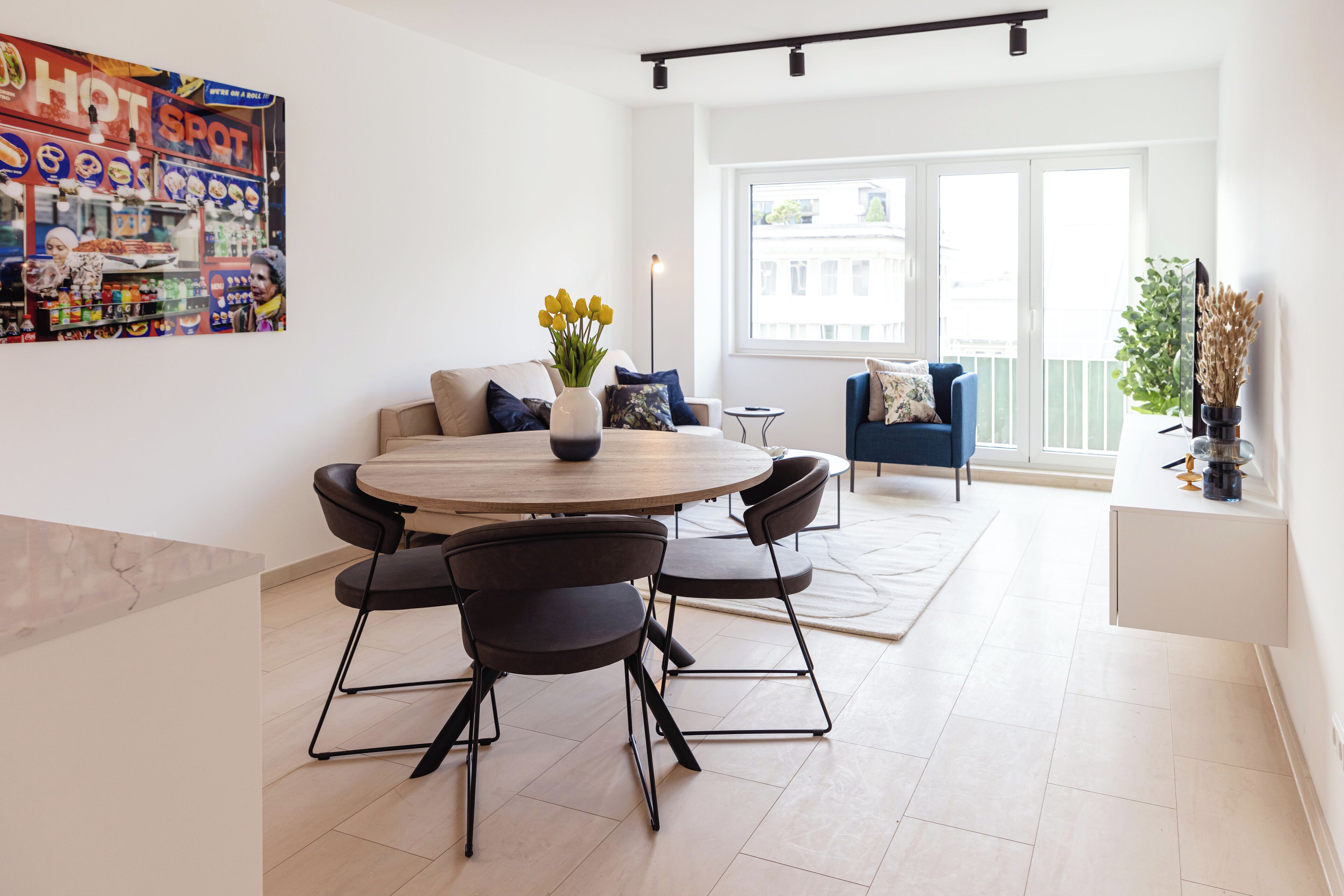 Una sala moderna con una mesa de comedor redonda, sillas negras, un sofá con cojines azules y un ventanal con vista a edificios.