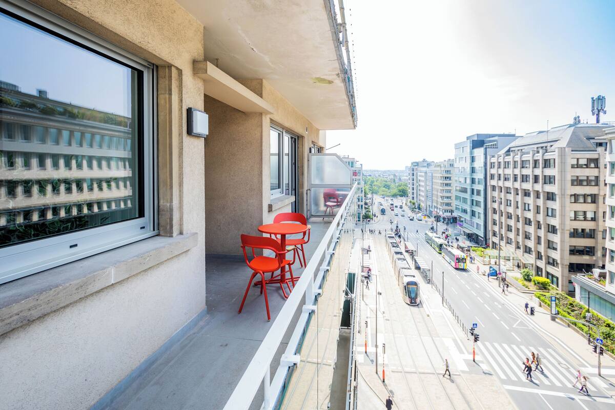 Un balcón con sillas rojas y un ventanal grande con vistas a una calle concurrida, edificios y un tranvía.