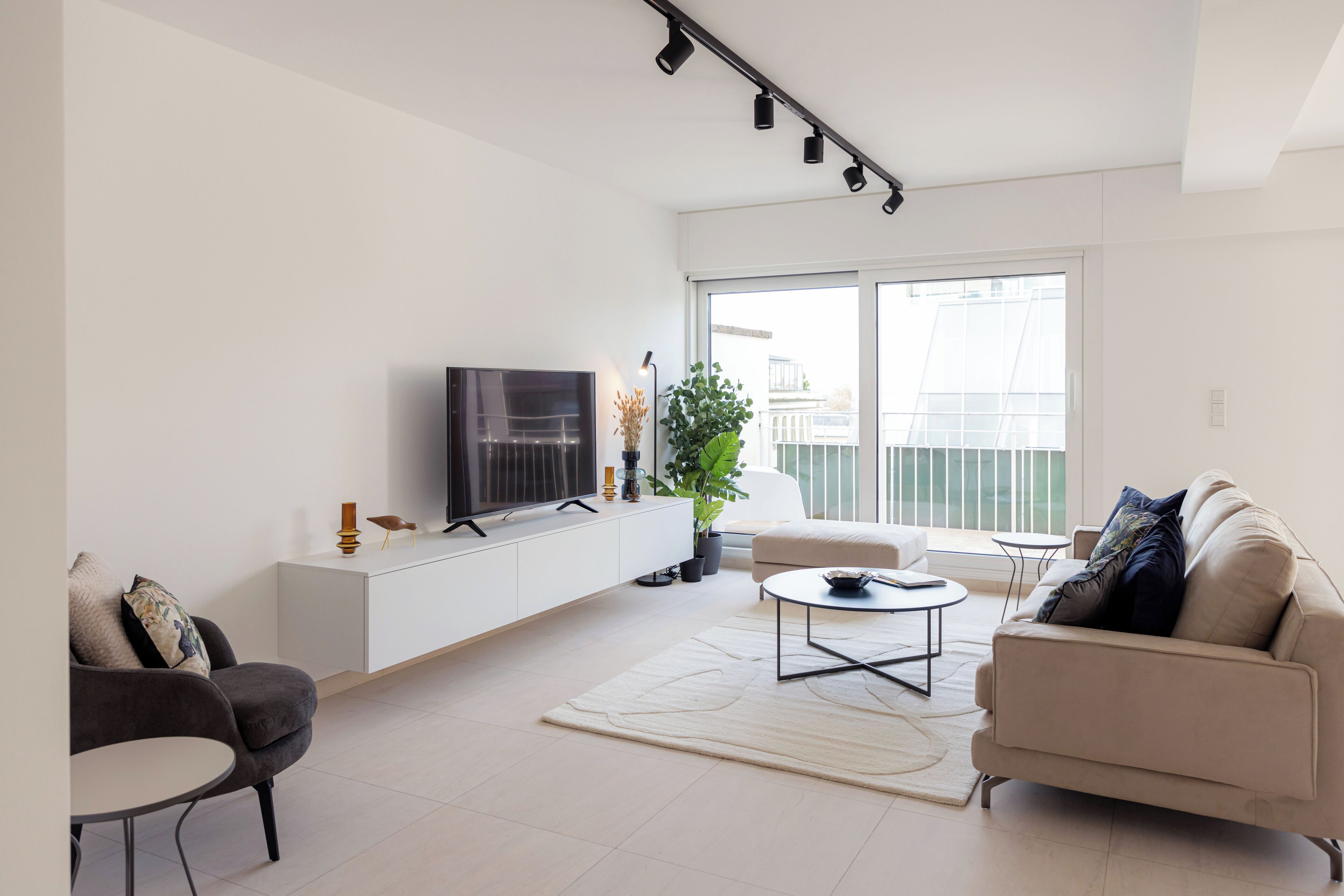 A modern living room with a flat-screen TV, a white sofa, a round coffee table, and a large window offering a view of buildings outside.