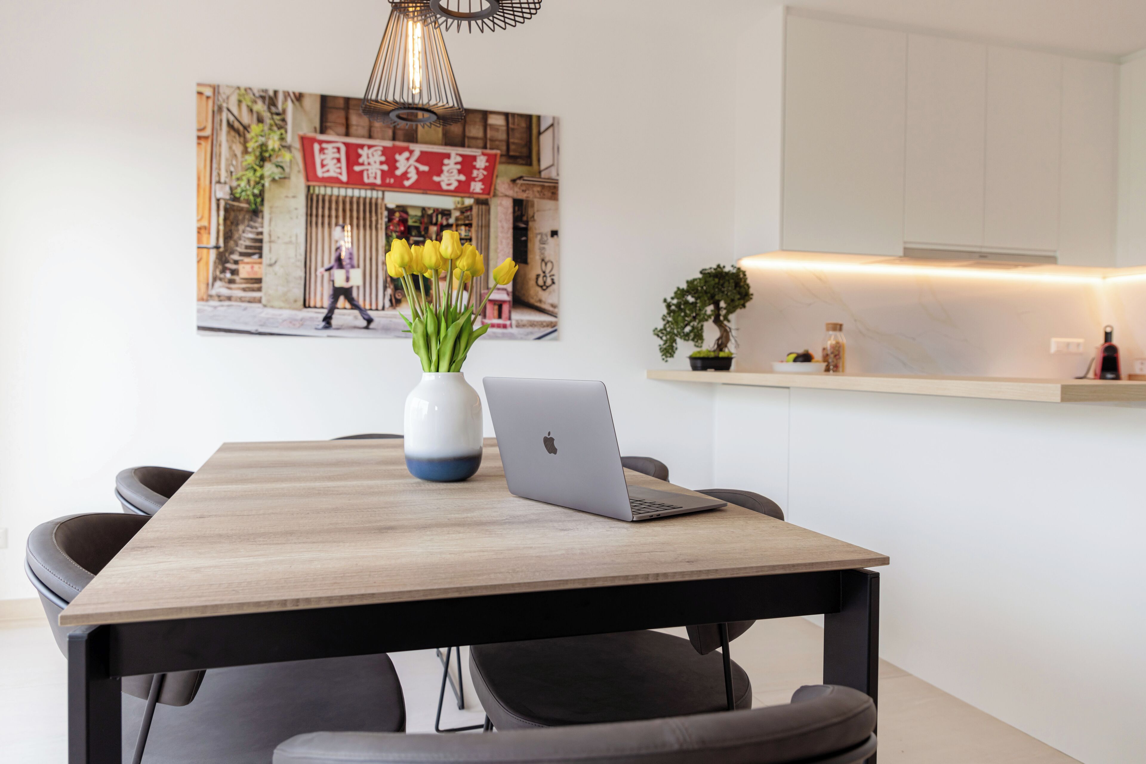 Uma zona de refeições moderna com uma mesa de madeira, cadeiras pretas, um laptop e um vaso de flores amarelas.