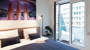 Une chambre d’hôtel moderne avec un grand lit, un ordinateur portable posé sur le lit, une plante en pot et une vue sur la ville depuis les portes du balcon.