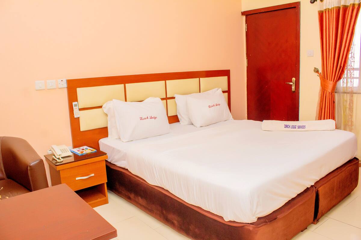 A hotel room with a wooden headboard, two white pillows, a bed with a white bedspread, a wooden nightstand with a phone and a book, and a red door.