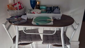 Un coin repas avec une table et des chaises en métal blanc, un panier en osier, un saladier orné de motifs floraux et un plateau avec une assiette verte et des couverts.