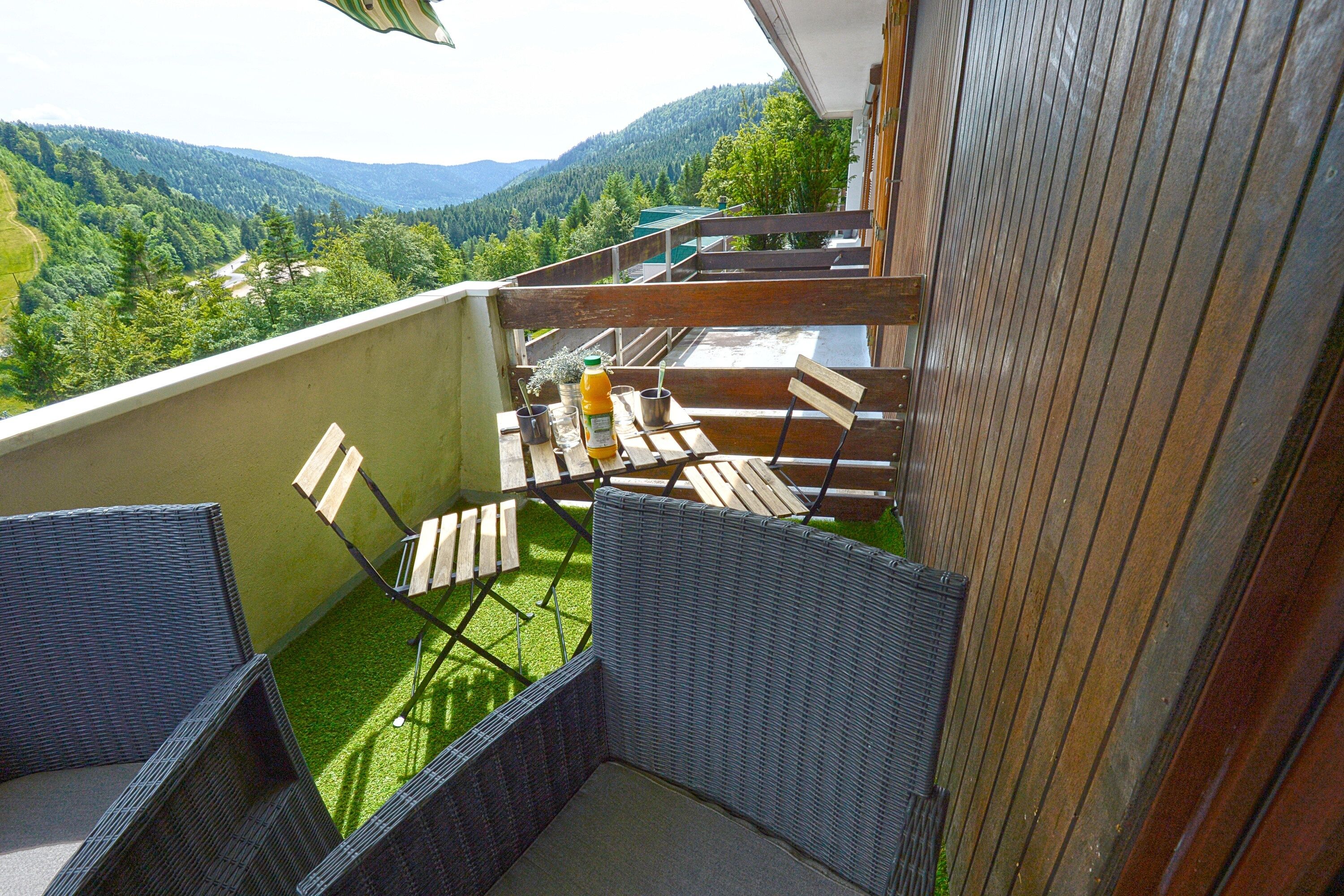 A balcony with wicker furniture, a table with a bottle and a glass, and a view of a forested valley.