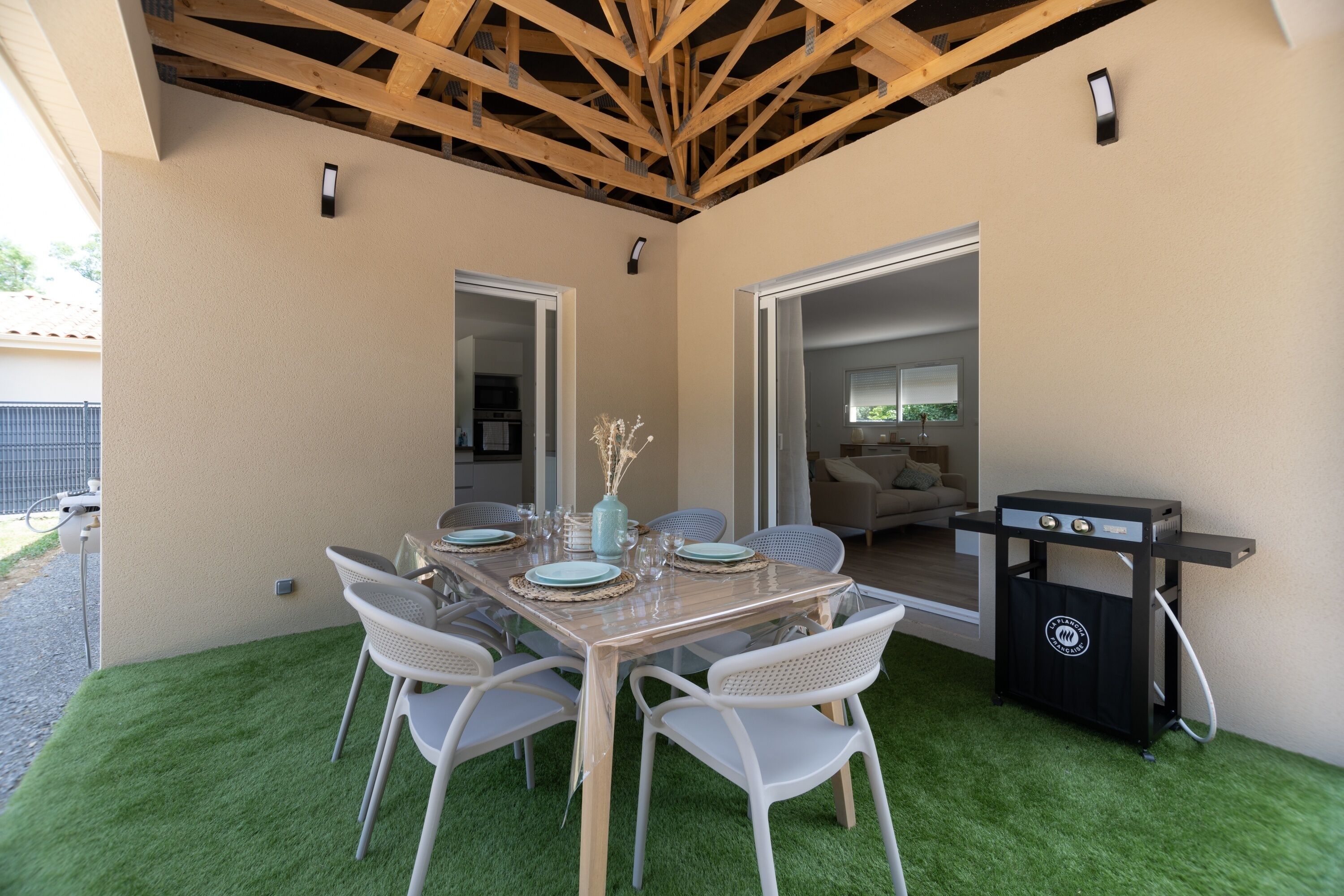 Een terras buiten met een houten tafel voor vier personen, omringd door witte stoelen, een barbecue en uitzicht op het huis.