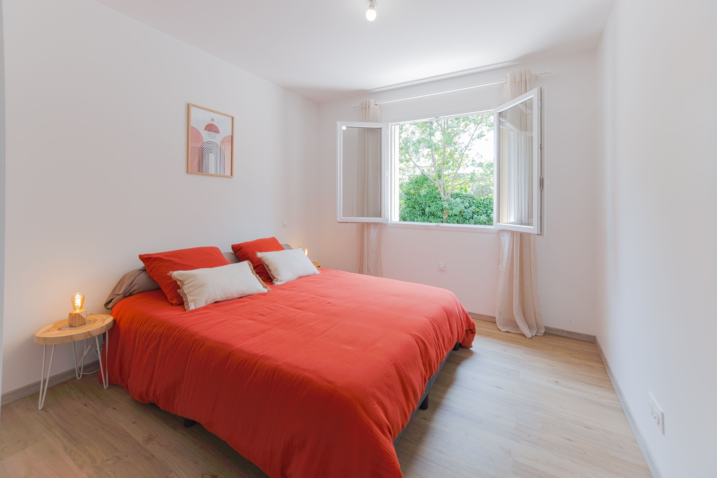 Een slaapkamer met een groot bed, een nachtkastje met een lamp, een raam met gordijnen en een lijst met een afbeelding aan de muur.