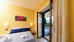 A hotel room with a bed, two pillows, a nightstand, a painting of a rose, and a view of a balcony with a hot tub. - B&B La Rosa (Ischia)