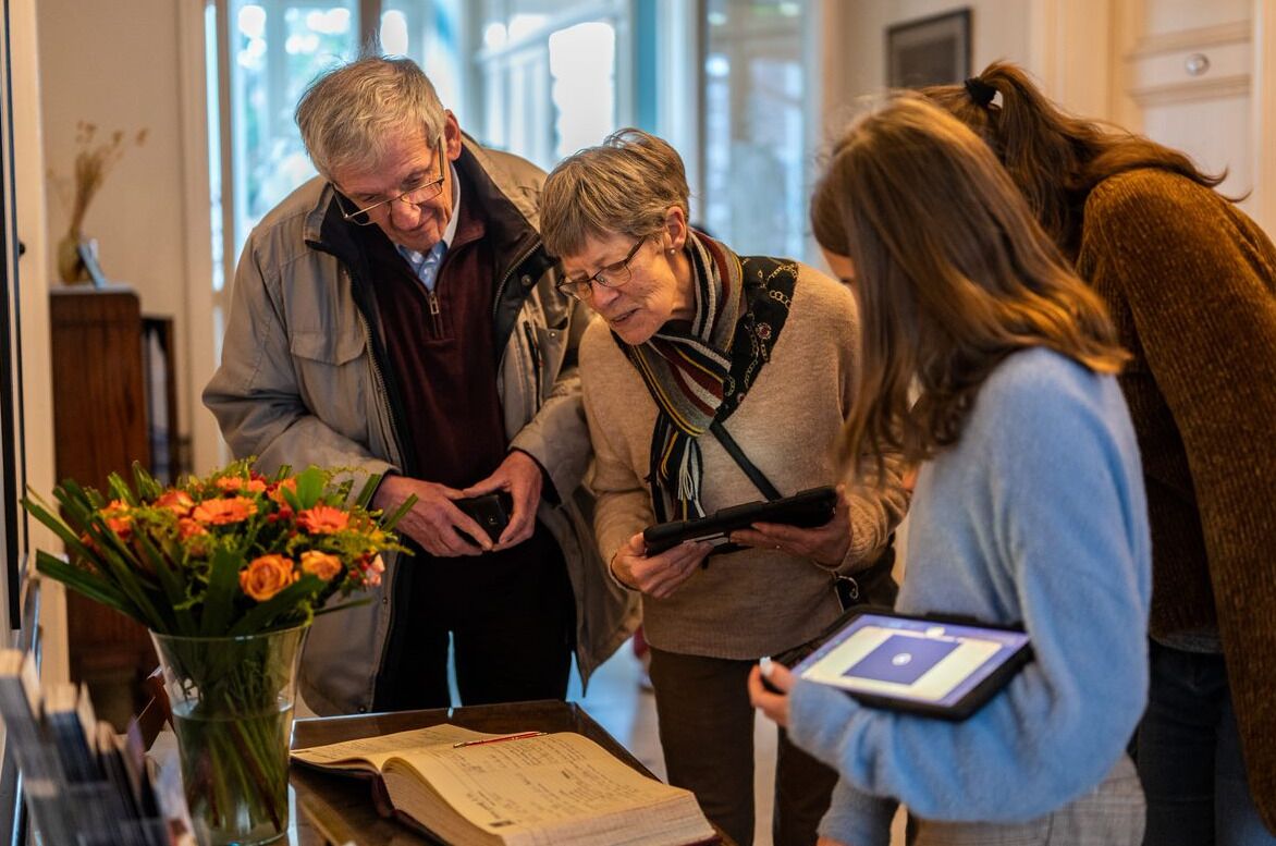 A group of people gathered around a table with a book and a tablet, in a room with a vase of flowers and a wooden cabinet.