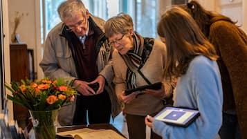 A group of people gathered around a table with a book and a tablet, in a room with a vase of flowers and a wooden cabinet.