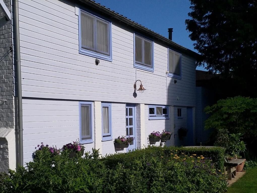 Een tweelaags gebouw met een witte gevel en blauwe accenten. Op de vensterbanken staan bloembakken en in de tuin een bank.