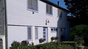 Een tweelaags gebouw met een witte gevel en blauwe accenten. Op de vensterbanken staan bloembakken en in de tuin een bank.