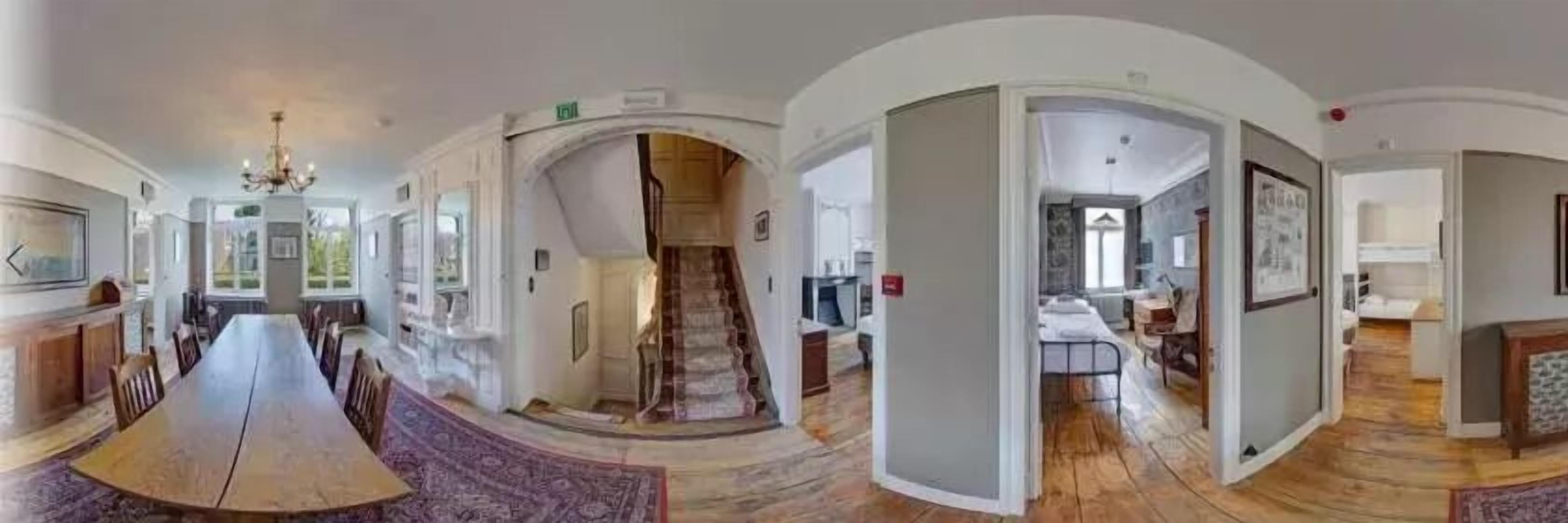 Vue panoramique d’un vaste espace de vie comprenant une grande table à manger en bois, un escalier et un coin cuisine.