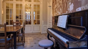 Ett piano med noter, ett matbord med stolar och ett stort, välvt fönster med glasrutor.