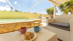 Zona piscina con una poltrona prendisole, un tavolo con un piatto di croissant e due tazze, e una piscina.