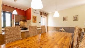 Zona pranzo con tavolo in legno e sedie in vimini, cucina con pareti rosse e soggiorno con divano e cuscini.