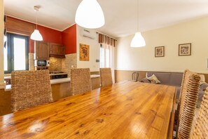 A dining area with a wooden table and woven chairs, a kitchen with red walls, and a living area with a sofa and cushions.