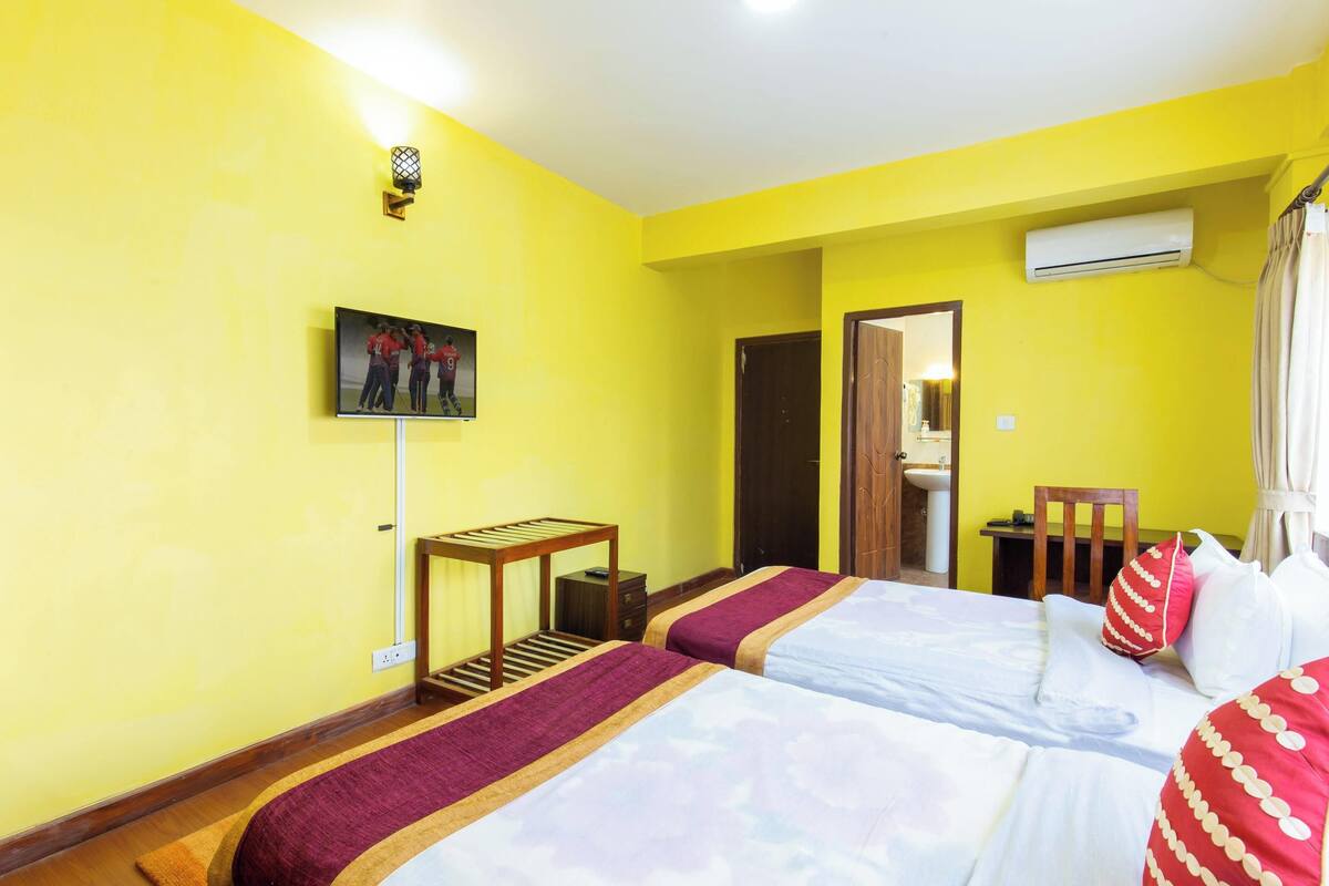 A hotel room with two beds, a TV mounted on the wall, a small table, and an air conditioning unit.